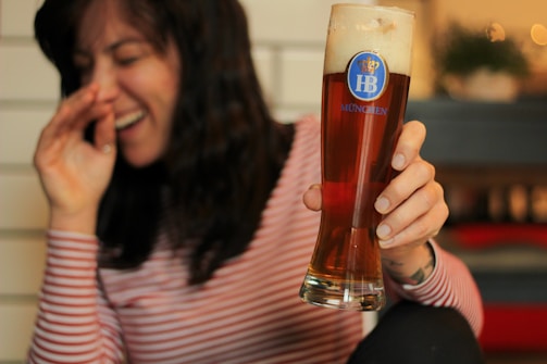 A person wearing a striped shirt is laughing while holding a tall glass of beer. The beer glass has a label with the text 'HB MÜNCHEN' and a crown symbol. The background appears blurred, with hints of warm lighting and indoor decorations.