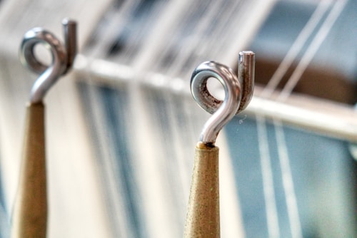 Close-up of two metal hooks with a spiral structure, positioned in the foreground with blurred vertical strings or threads in the background. The hooks are attached to wooden rods, with a metallic sheen contributing to the industrial feel of the image.