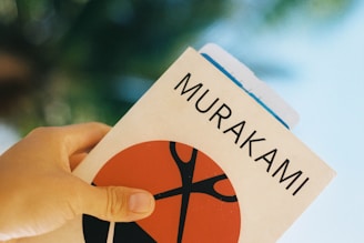 A hand holding a book with the name Murakami on the cover. The cover features a minimalist design with a large red and black graphic element. The background is blurred, giving a soft focus effect.
