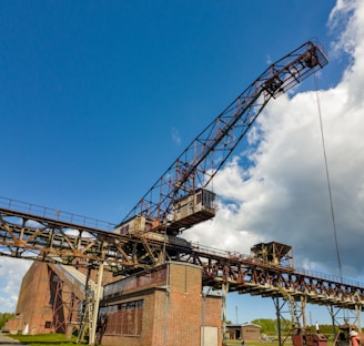 A large industrial crane with a steel framework towering in the foreground under a bright blue sky scattered with some clouds. The structure is a mix of red and rusted steel parts, and there is a brick building at its base.