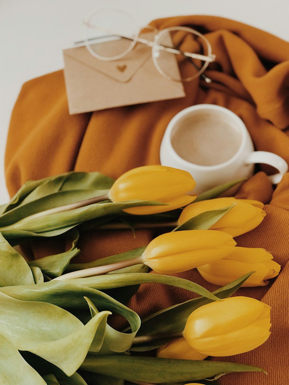 yellow tulip flowers near white ceramic mug, brown envelope, and gold framed eyeglasses
