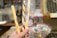 Three colorful candy sticks with yellow, white, and brown stripes are being held in a hand. The background shows a blurred confectionery display with various bags of candies and treats.