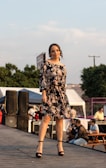 A woman is standing confidently on an outdoor stage wearing a black floral dress and black heels. Around her, there are people sitting at picnic tables and equipment like speakers and tents in the background. The scene suggests an event or fashion show with a relaxed, open-air setting.