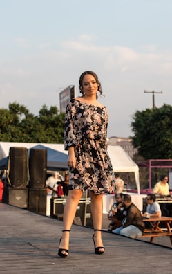 A woman is standing confidently on an outdoor stage wearing a black floral dress and black heels. Around her, there are people sitting at picnic tables and equipment like speakers and tents in the background. The scene suggests an event or fashion show with a relaxed, open-air setting.