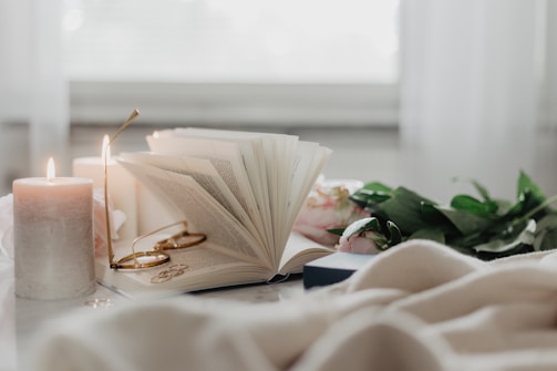 white book laying on table, pages opens with glasses sat in the inside. lit candles are beside it.