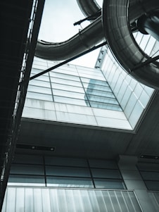 A modern architectural structure with large metal pipes and sleek glass panels viewed from a low angle. The design incorporates industrial elements with a futuristic style, featuring reflections and a gray-blue color palette.