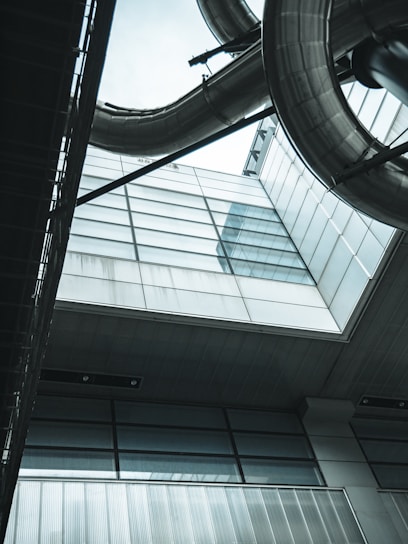 A modern architectural structure with large metal pipes and sleek glass panels viewed from a low angle. The design incorporates industrial elements with a futuristic style, featuring reflections and a gray-blue color palette.