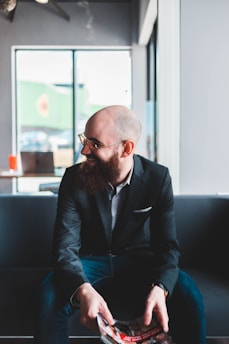 A man with a beard is sitting on a couch in a modern setting, holding a magazine. He is wearing glasses and a dark blazer over a white shirt. Soft natural light filters through large windows behind him, creating a relaxed atmosphere.