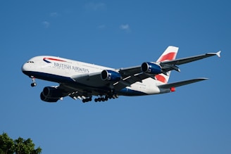 A British Airways airplane is flying against a clear blue sky. The aircraft appears to be in mid-flight, showcasing its landing gear and engines prominently. The sunlight reflects off the body, highlighting the airline's branding and color scheme. Small clouds can be seen scattered in the sky.