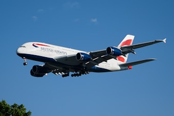 A British Airways airplane is flying against a clear blue sky. The aircraft appears to be in mid-flight, showcasing its landing gear and engines prominently. The sunlight reflects off the body, highlighting the airline's branding and color scheme. Small clouds can be seen scattered in the sky.