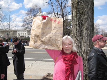A woman stands beside a tree on a sidewalk, holding up a handmade sign advocating for a vote. She is wearing a pink jacket and red scarf. Behind her, a few people stand near a road with a significant building in the background, likely a government structure. The sky is partly cloudy.