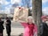 A woman stands beside a tree on a sidewalk, holding up a handmade sign advocating for a vote. She is wearing a pink jacket and red scarf. Behind her, a few people stand near a road with a significant building in the background, likely a government structure. The sky is partly cloudy.