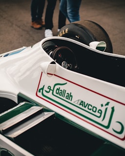 A vintage racing car with white and green livery is prominently displayed, featuring Arabic script and logos on the body. Part of a tire and a few people's legs in jeans and shoes are visible in the background.