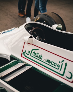 A vintage racing car with white and green livery is prominently displayed, featuring Arabic script and logos on the body. Part of a tire and a few people's legs in jeans and shoes are visible in the background.