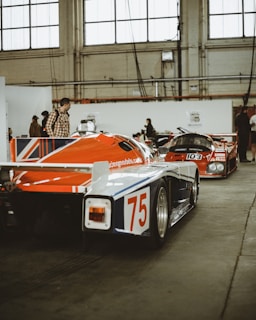 Two vintage racing cars are parked inside what appears to be an industrial or exhibition space with high ceilings and large windows. The cars are painted with vibrant livery, featuring red, white, and blue colors. People are walking around and observing the vehicles.