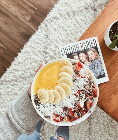 A bowl filled with a colorful smoothie topped with sliced bananas, shredded coconut, chia seeds, and strawberries. The bowl is held by a person wearing a gray sweater with jeans visible below. A magazine with three women on the cover and the title 'Edition Papier' is on a wooden table nearby, next to a small potted plant. The background includes a light, textured rug and wooden floor.