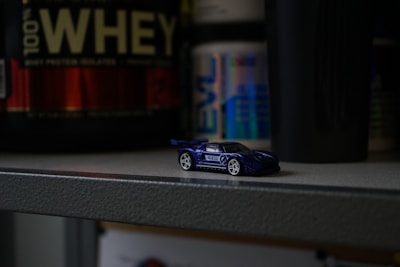 A small toy race car in blue and white sits on a gray shelf with containers of protein powder and supplements in the background. The setting appears dimly lit, creating a contrasting focus on the toy car.
