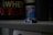 A small toy race car in blue and white sits on a gray shelf with containers of protein powder and supplements in the background. The setting appears dimly lit, creating a contrasting focus on the toy car.