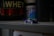 A small toy race car in blue and white sits on a gray shelf with containers of protein powder and supplements in the background. The setting appears dimly lit, creating a contrasting focus on the toy car.