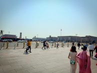 People are walking in an open, paved area with a wide expanse of sky. In the background, there is a large flagpole with a red flag and a grand, official-looking building. Vehicles and a red bus are seen on the road beyond a low decorative fence.