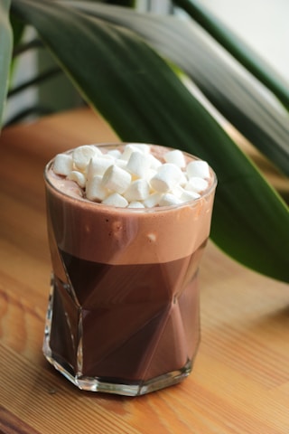 A glass filled with rich chocolate milk is topped with a generous amount of mini marshmallows. The drink sits on a wooden surface with a background of large, leafy green plants, adding a fresh and natural ambiance.