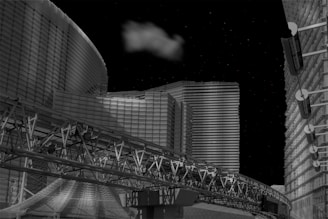 A futuristic cityscape with modern, geometric buildings illuminated against a night sky. A cloud floats above, adding contrast to the metallic and glass structures. An intricate transport track runs across the foreground, contributing to the architectural complexity.