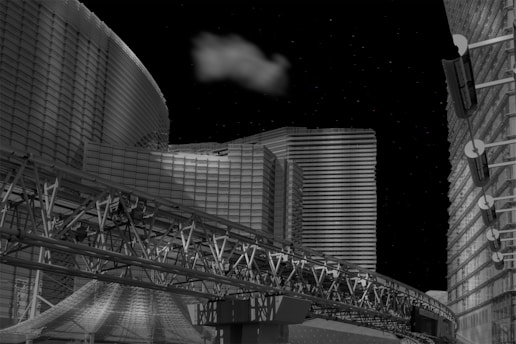 A futuristic cityscape with modern, geometric buildings illuminated against a night sky. A cloud floats above, adding contrast to the metallic and glass structures. An intricate transport track runs across the foreground, contributing to the architectural complexity.