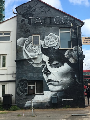 A detailed black and white mural on a building depicts a woman with a skull and floral design. The word 'TATTOO' is prominently displayed at the top. The mural incorporates elements of roses and decorative swirls around the central figure. Adjacent signage indicates tattoo removal services.