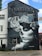 A detailed black and white mural on a building depicts a woman with a skull and floral design. The word 'TATTOO' is prominently displayed at the top. The mural incorporates elements of roses and decorative swirls around the central figure. Adjacent signage indicates tattoo removal services.