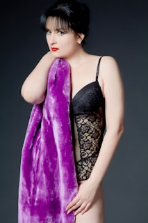 A woman with dark hair and red lipstick is wearing a black lace bodysuit and holding a plush purple fabric. The background is plain and dark, contrasting with her outfit and the fabric.