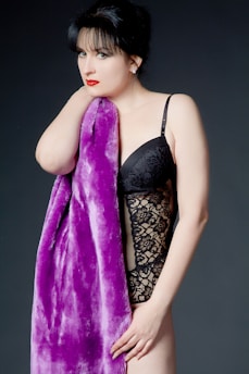 A woman with dark hair and red lipstick is wearing a black lace bodysuit and holding a plush purple fabric. The background is plain and dark, contrasting with her outfit and the fabric.