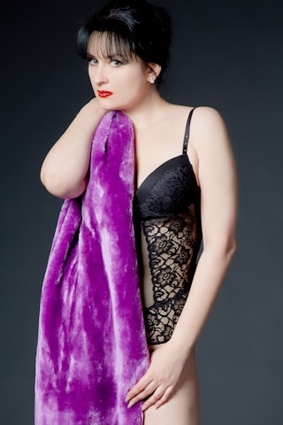 A woman with dark hair and red lipstick is wearing a black lace bodysuit and holding a plush purple fabric. The background is plain and dark, contrasting with her outfit and the fabric.