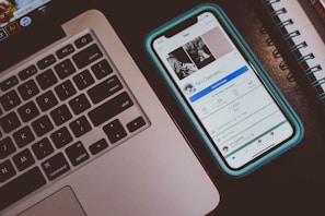A smartphone displaying a social media profile page lies next to a laptop keyboard. The phone is in a blue case, and the profile page includes a cover photo with three images. Part of a spiral notebook is visible to the side.