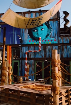 A vibrant mural of a child's face painted on a corrugated metal wall with colorful hearts below. The setting includes ropes tied around wooden posts, resembling a nautical or pirate theme. There are large tarps with knots hanging overhead. The building is adorned with industrial and urban elements, including a striped pole and a rustic staircase.