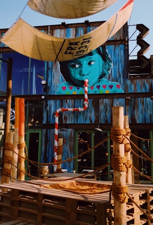 A vibrant mural of a child's face painted on a corrugated metal wall with colorful hearts below. The setting includes ropes tied around wooden posts, resembling a nautical or pirate theme. There are large tarps with knots hanging overhead. The building is adorned with industrial and urban elements, including a striped pole and a rustic staircase.