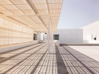 A modern architectural scene featuring clean, geometric lines and a grid-like shadow pattern cast on the floor. The space is dominated by light, neutral tones, with an overhang providing shade and an open feel. The structure showcases a minimalist design with a focus on symmetry and texture.