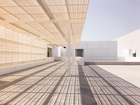 A modern architectural scene featuring clean, geometric lines and a grid-like shadow pattern cast on the floor. The space is dominated by light, neutral tones, with an overhang providing shade and an open feel. The structure showcases a minimalist design with a focus on symmetry and texture.