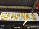 A wooden sign with the word 'BANANAS' in large yellow letters. The sign is hanging from a wooden structure with a corrugated metal roof. Below the sign, there are other signs and decor pieces, including one that says 'GUARAPO' and another indicating 'WORLD FAMOUS'.