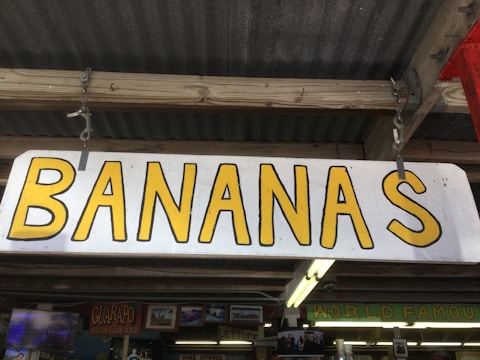 A wooden sign with the word 'BANANAS' in large yellow letters. The sign is hanging from a wooden structure with a corrugated metal roof. Below the sign, there are other signs and decor pieces, including one that says 'GUARAPO' and another indicating 'WORLD FAMOUS'.