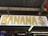 A wooden sign with the word 'BANANAS' in large yellow letters. The sign is hanging from a wooden structure with a corrugated metal roof. Below the sign, there are other signs and decor pieces, including one that says 'GUARAPO' and another indicating 'WORLD FAMOUS'.