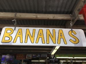 A wooden sign with the word 'BANANAS' in large yellow letters. The sign is hanging from a wooden structure with a corrugated metal roof. Below the sign, there are other signs and decor pieces, including one that says 'GUARAPO' and another indicating 'WORLD FAMOUS'.
