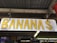 A wooden sign with the word 'BANANAS' in large yellow letters. The sign is hanging from a wooden structure with a corrugated metal roof. Below the sign, there are other signs and decor pieces, including one that says 'GUARAPO' and another indicating 'WORLD FAMOUS'.
