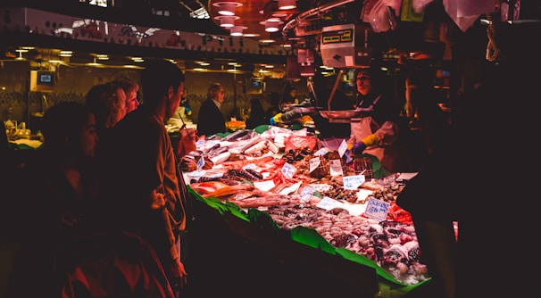 A bustling indoor fish market with customers examining various seafood displayed on a brightly lit counter. The scene is lit with warm, overhead lighting, enhancing the vibrant colors of the seafood. Several people, including a vendor, are visible, engaging in what appears to be a transaction.
