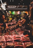 A bustling market scene with several people interacting around a stall filled with various packaged goods. Colorful banners hang above the stall, adding a vibrant atmosphere. The lighting is subdued with a warm glow, highlighting the array of products on display.