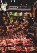 A bustling market scene with several people interacting around a stall filled with various packaged goods. Colorful banners hang above the stall, adding a vibrant atmosphere. The lighting is subdued with a warm glow, highlighting the array of products on display.