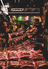 A bustling market scene with several people interacting around a stall filled with various packaged goods. Colorful banners hang above the stall, adding a vibrant atmosphere. The lighting is subdued with a warm glow, highlighting the array of products on display.