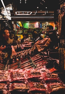 A bustling market scene with several people interacting around a stall filled with various packaged goods. Colorful banners hang above the stall, adding a vibrant atmosphere. The lighting is subdued with a warm glow, highlighting the array of products on display.