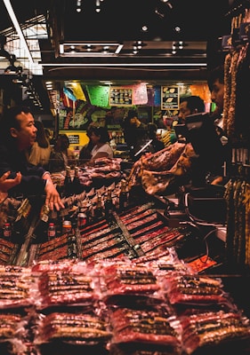 A bustling market scene with several people interacting around a stall filled with various packaged goods. Colorful banners hang above the stall, adding a vibrant atmosphere. The lighting is subdued with a warm glow, highlighting the array of products on display.