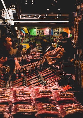 A bustling market scene with several people interacting around a stall filled with various packaged goods. Colorful banners hang above the stall, adding a vibrant atmosphere. The lighting is subdued with a warm glow, highlighting the array of products on display.