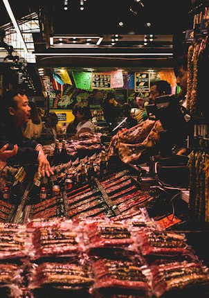 A bustling market scene with several people interacting around a stall filled with various packaged goods. Colorful banners hang above the stall, adding a vibrant atmosphere. The lighting is subdued with a warm glow, highlighting the array of products on display.